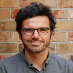 Porträtfoto Johannes Rupp mit dunklem Haar, Brille und grauem Hemd lächelt in die Kamera.