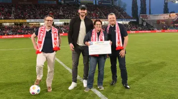 Vier Personen stehen im Stadion von Energie Cottbus auf dem Rasen und halten einen Scheck hoch