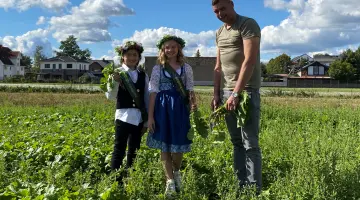 Die Rübchenprinzessin und -prinz stehen bei Sonnenschein mit dem Bauern auf dem Rübchenfeld und lächeln.
