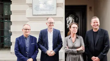 Vier Personen stehen von einer Berliner Gedenktafel, die an einer Hauswand angebracht ist