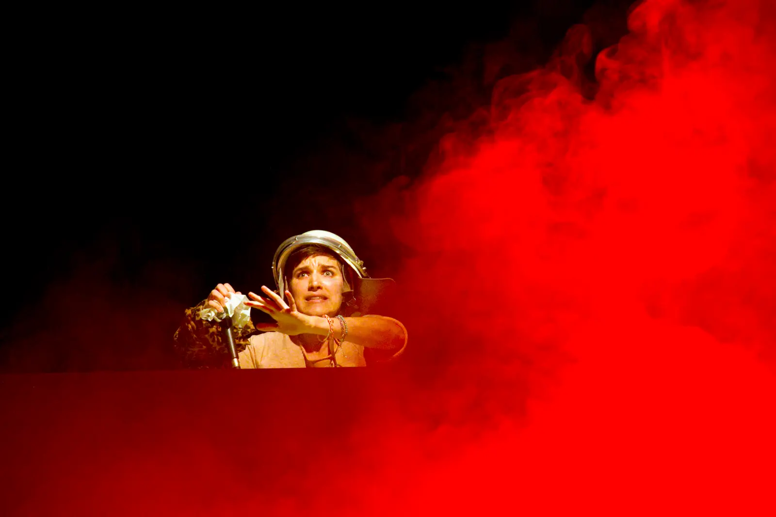 Person mit Helm auf einer Bühne erscheint aus rotem Nebel und hält ein Mikrofon, dramatisch beleuchtet vor schwarzem Hintergrund.