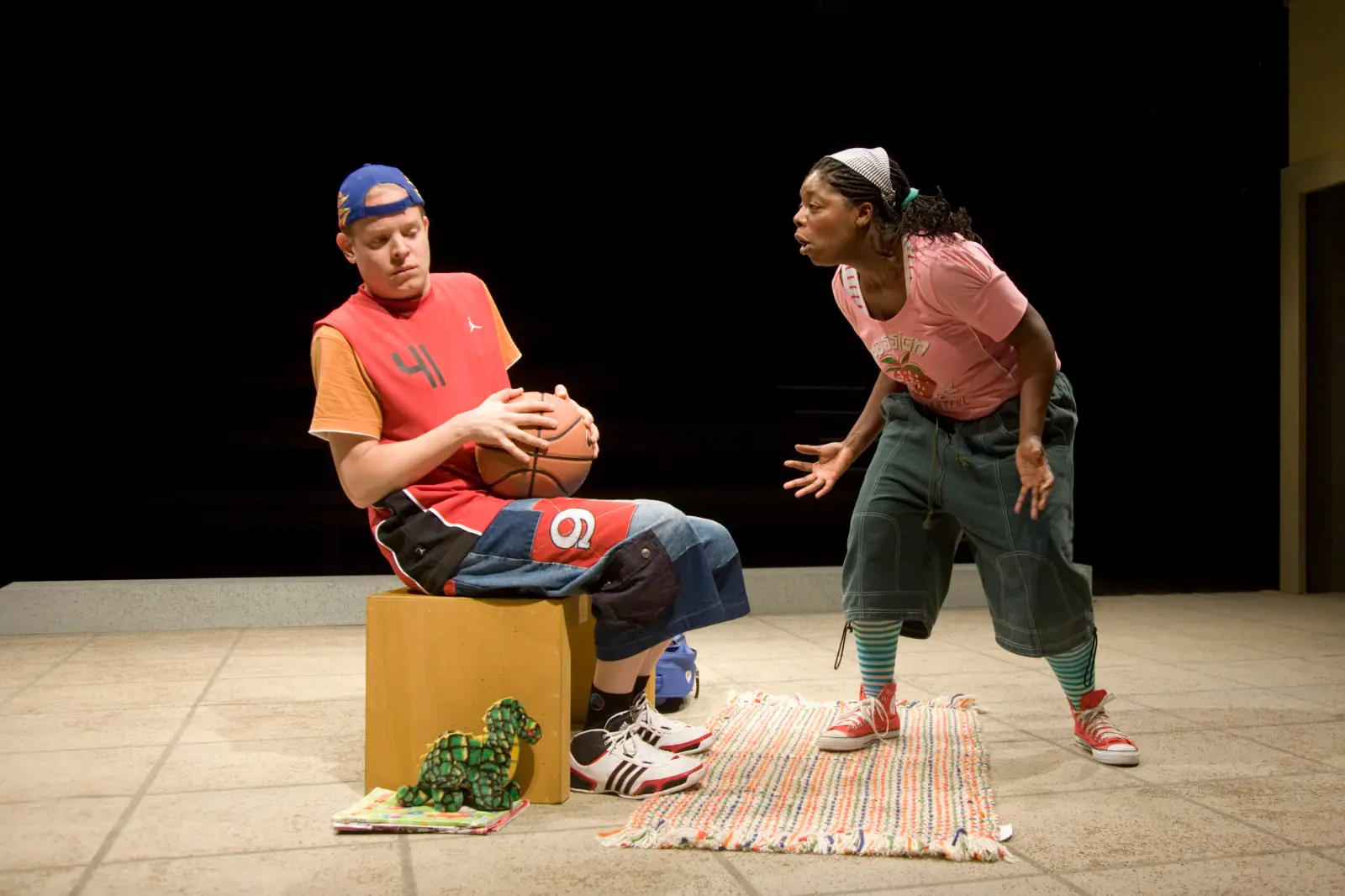 Zwei Personen auf einer Bühne. Eine Person mit Basketball sitzt eingeschüchtert auf einem Hocker, die andere steht daneben und redet auf sie ein.