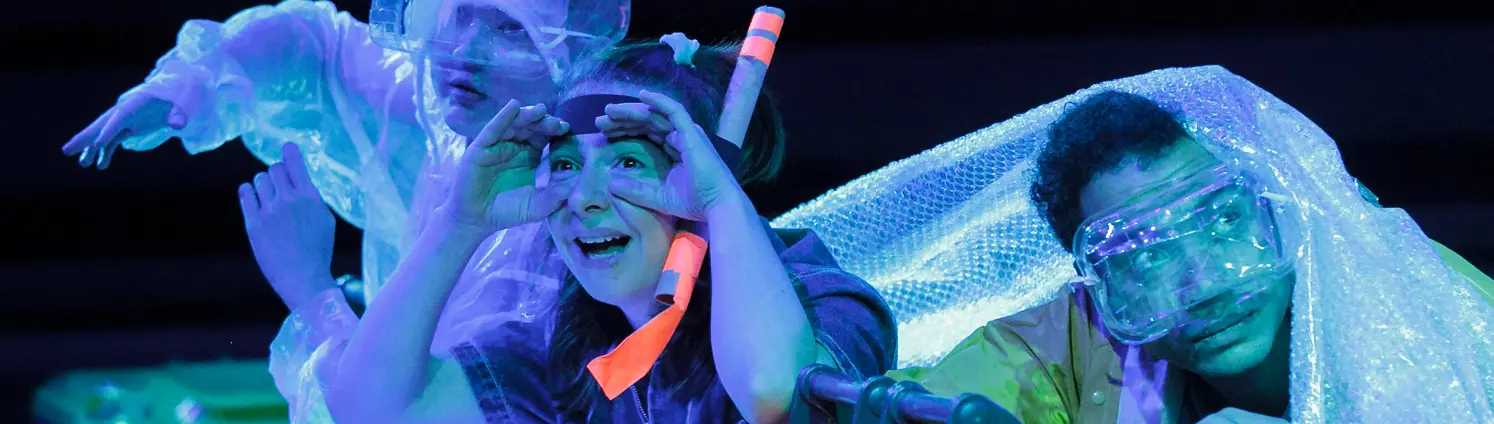 Drei Personen in durchsichtiger Schutzkleidung sitzen auf einer Bühne bei blauem Licht, umgeben von glitzernden Materialien und Requisiten.