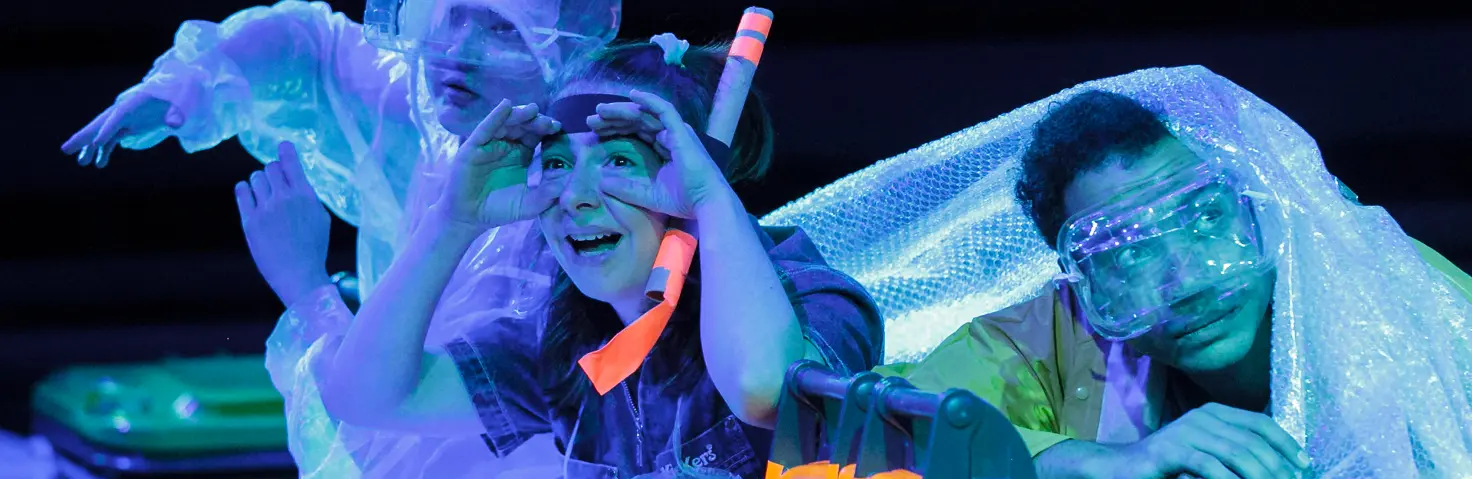 Drei Personen in durchsichtiger Schutzkleidung sitzen auf einer Bühne bei blauem Licht, umgeben von glitzernden Materialien und Requisiten.