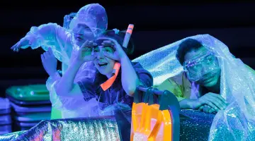 Drei Personen in durchsichtiger Schutzkleidung sitzen auf einer Bühne bei blauem Licht, umgeben von glitzernden Materialien und Requisiten.