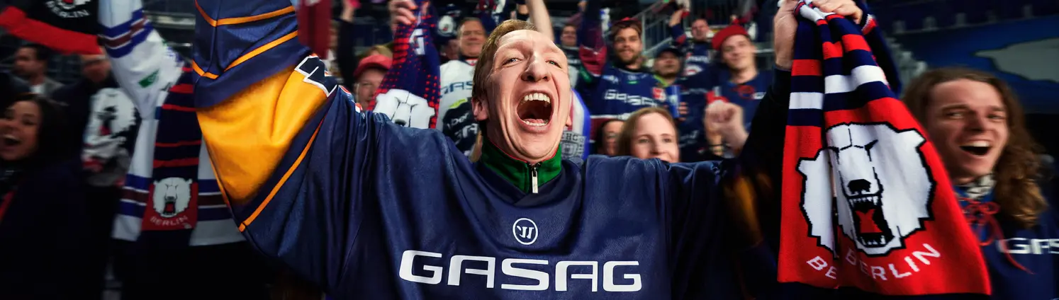 Eishockey-Fans mit Schals jubeln im Stadion, Person im Vordergrund mit erhobenem Arm und Logo der Eisbären Berlin auf dem Trikot.