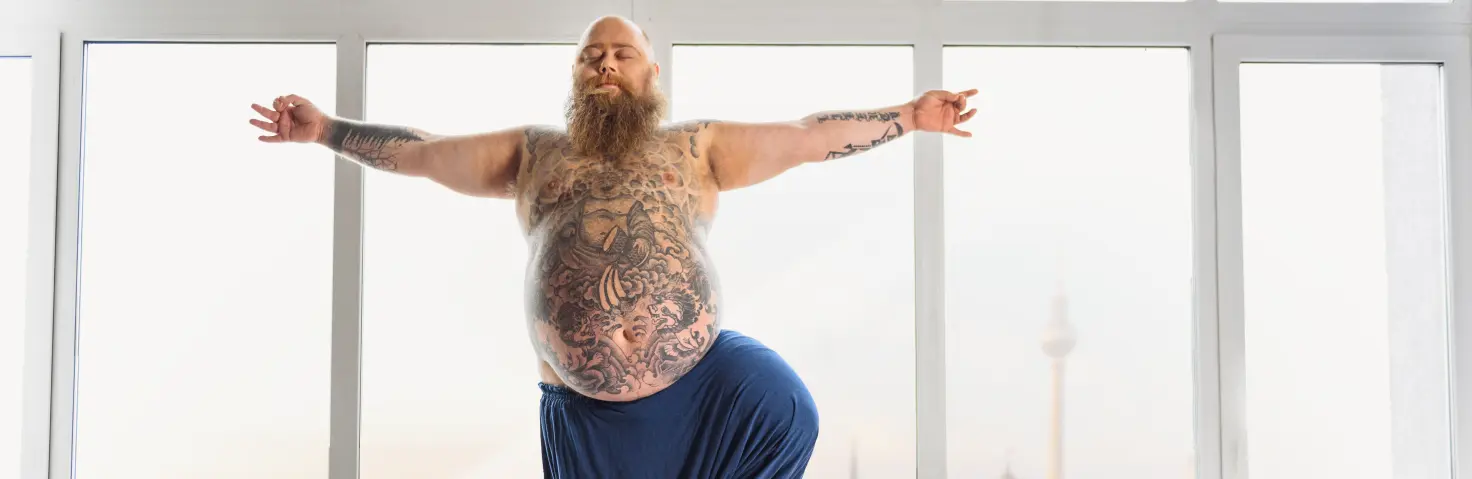 Ein beleibter Mann in blauer Stoffhose steht von einem Fenster mit Berlin-Skyline und macht Yoga.