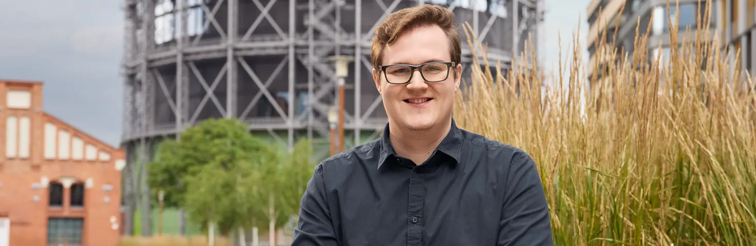 Ein Auszubildender zum Kaufmann für Digitalisierungsmanagement hält ein Handy in der Hand. Hinter ihm der Gasometer auf dem EUREF-Campus.