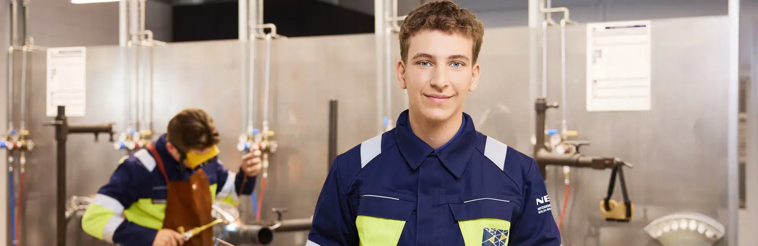 Ein lächelnder Auszubildender der NBB Netzgesellschaft in Arbeitskleidung hält eine Schutzbrille und zwei dünne Rohrleitungen in den Händen.