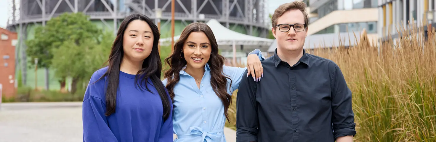 Drei Auszubildende der NBB stehen nebeneinander vor dem Hauptsitz der GASAG-Gruppe in Berlin-Schöneberg und lächeln.