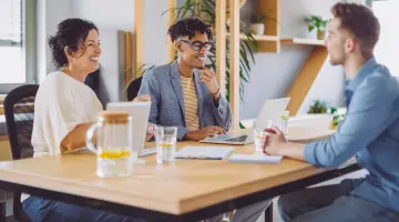 Zwei Menschen führen ein Bewerbungsgespräch mit einer Person, sitzen um einen Tisch herum und lachen freundlich