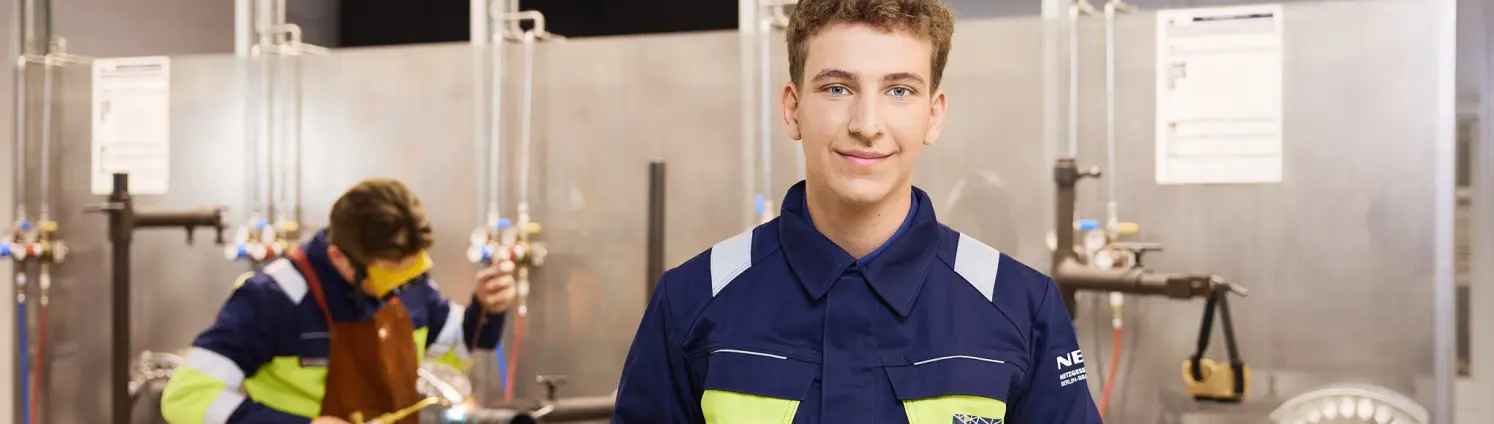 Person in Arbeitskleidung hält ein Prüfgerät in einer Werkstatt. Im Hintergrund werden Metallrohre mit Brennern bearbeitet.