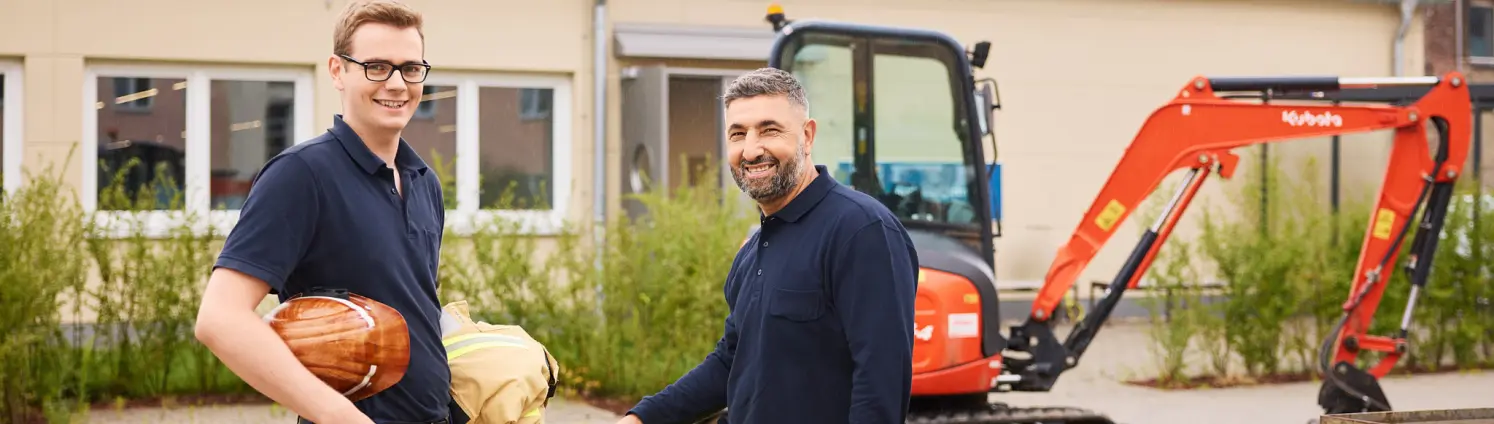 Zwei Mitarbeitende in Arbeitskleidung stehen auf einer Baustelle neben einer großen Rohrleitung, im Hintergrund ein Bagger und ein Betriebsgebäude.