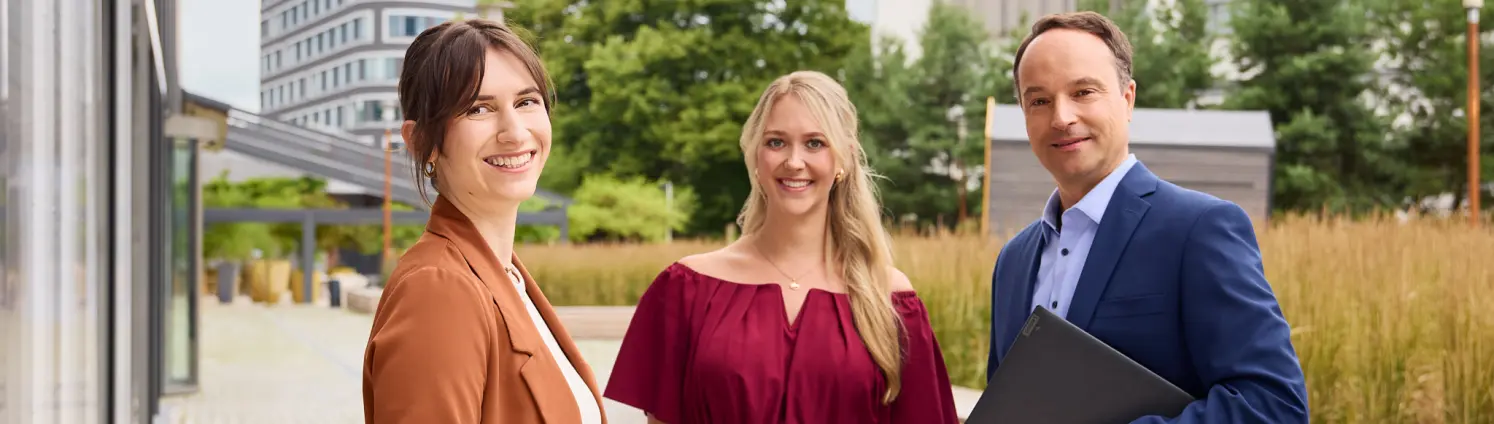 Drei Personen in Businesskleidung stehen im Außenbereich eines modernen Büroensembles. Eine Person hält eine Mappe, im Hintergrund Gebäude und Grün.