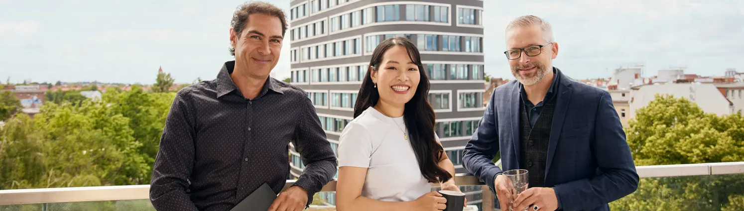 Drei Personen stehen auf einer Dachterrasse mit Blick über die Stadt, halten Getränke und Unterlagen. Im Hintergrund ein Gebäude und grüne Bäume.
