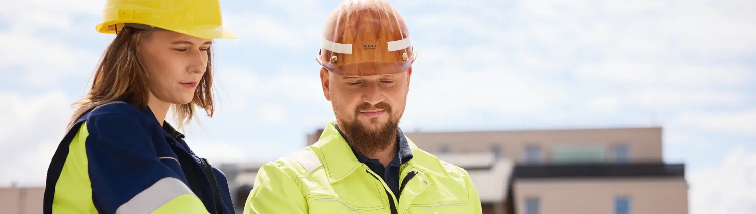 Zwei Personen in Warnschutzkleidung und Schutzhelm besprechen einen Bauplan auf einer Baustelle, im Hintergrund Gebäude und Himmel.