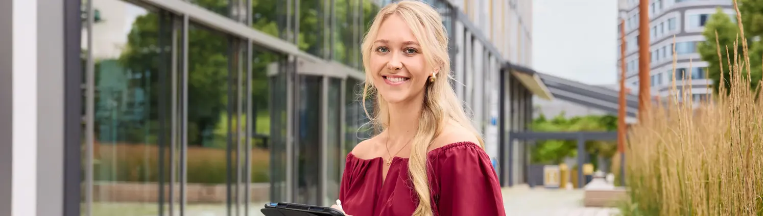 Person sitzt auf einer Holzbank vor einem modernen Glasgebäude und hält ein Tablet, umgeben von bepflanzten Außenflächen und urbaner Architektur.