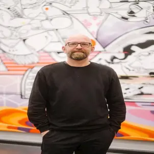 Ein Mann in schwarzer Kleidung, die Hände in den Hosentaschen, und steht vor einer bunt bemalten Wand