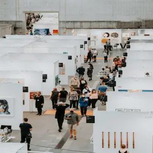 Besucher der Berlin Art Fair laufen einen Messegang entlang, links und rechts weiße Boxen, in denen Bilder ausgestellt sind.
