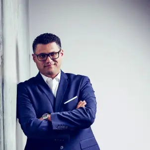 Herr Serdar Savas steht im Anzug gegen eine Wand gelehnt und lächelt