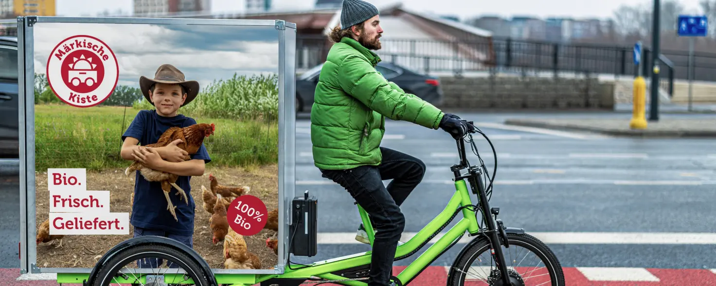 Lieferant auf Elektro-Lastenfahrrad mit Bio‑Lebensmittel‑Werbung der Märkischen Kiste überquert einen Zebrastreifen.