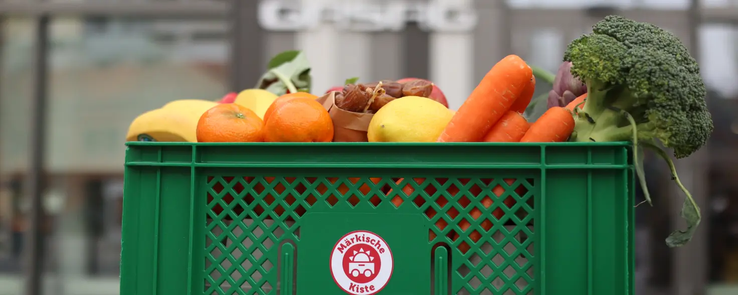 Grüne Lebensmittelkiste mit frischem Obst und Gemüse wie Karotten, Brokkoli, Äpfeln und Zitrusfrüchten vor einem Gebäude mit GASAG‑Schriftzug.