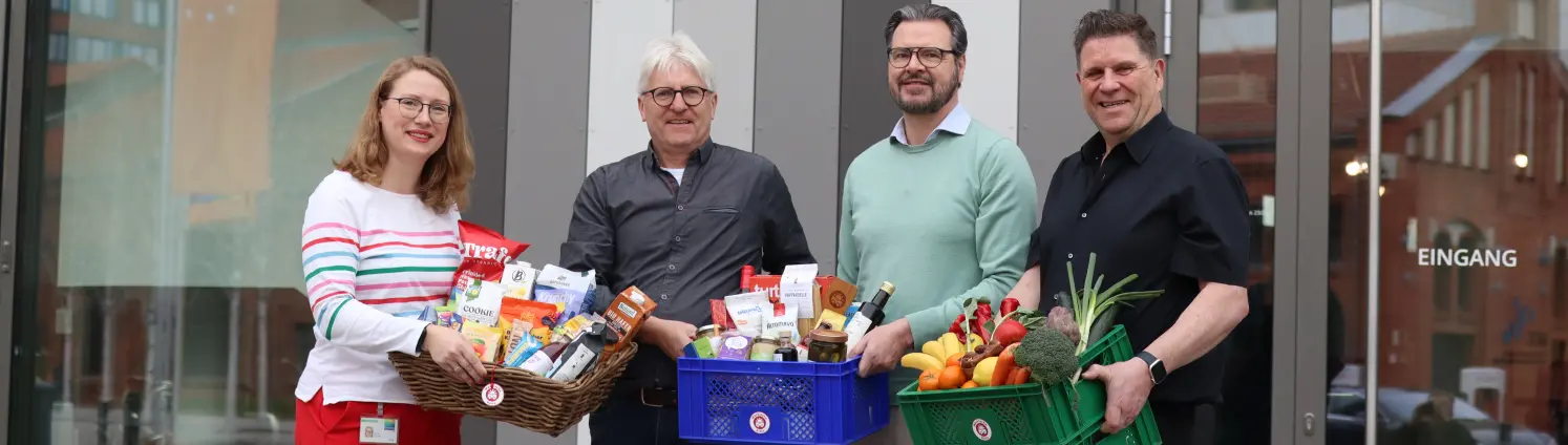 Vier Personen stehen vor dem Eingang eines Gebäudes mit der Aufschrift ‚GASAG‘ und halten Kisten mit Lebensmitteln und frischem Obst und Gemüse.