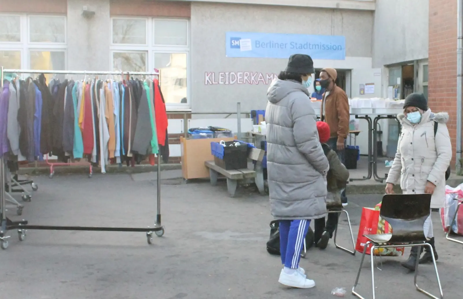 Links steht ein Kleiderständer mit Kleidung, auf der rechten Seite steht ein Mann in grauem Daunenmantel und blauer Hose