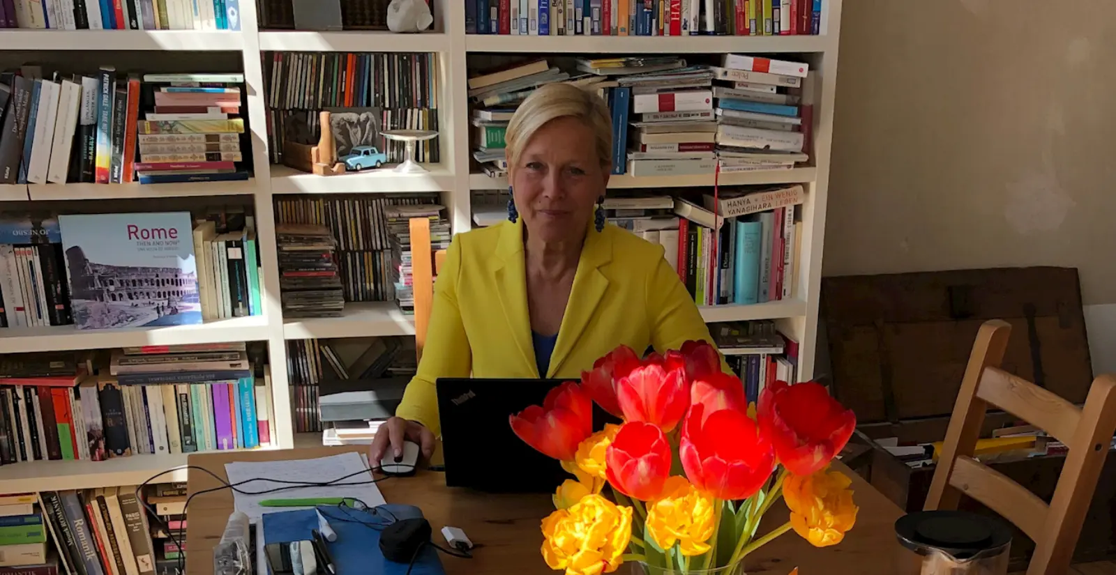 Birgit Jammes sitzt an einem Holztisch, vor ihr sind ein Laptop und ein Strauß Tulpen. Hinter ihr ist ein weißes Bücherregal.