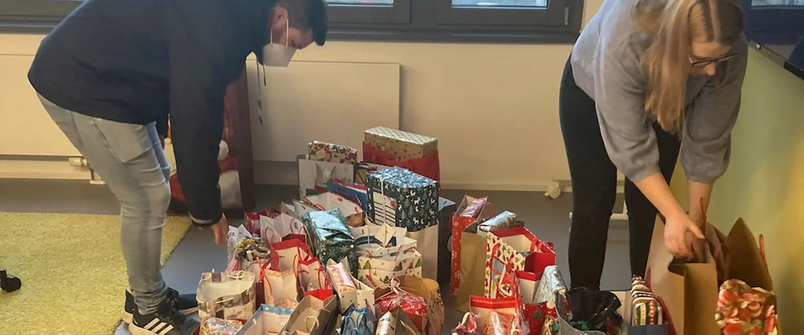 Auf dem Boden stehen einige Geschenktüten. Links steht ein Mann, rechts eine Frau und beide greifen nach einer Geschenktüte