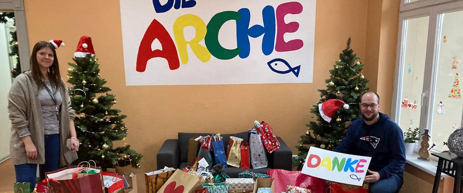 Zu beiden Seiten stehen Weihnachtsbäume, link steht eine Frau, rechts sitzt ein Mann mit einem 
