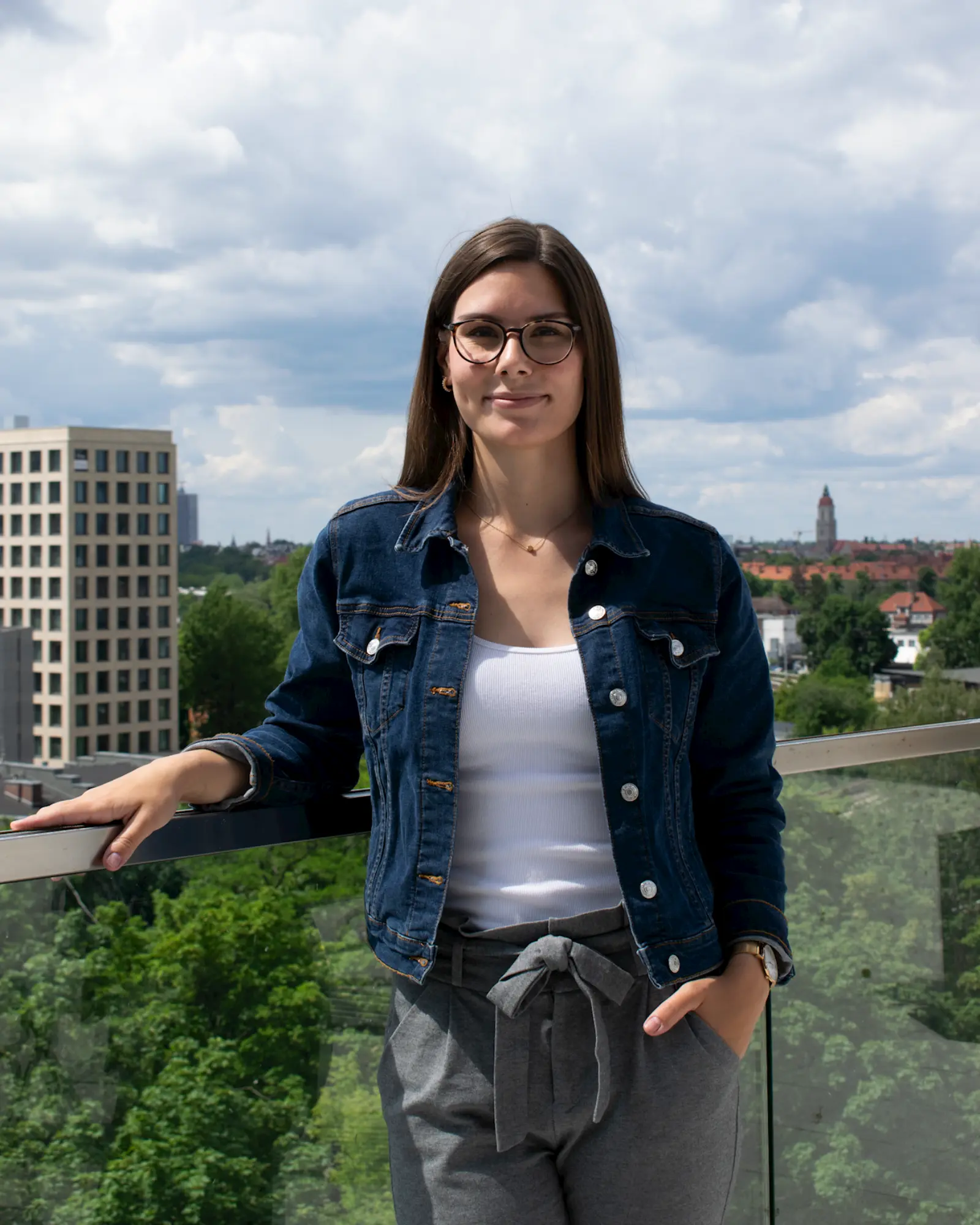 Lilly steht auf dem Balkon, ihr rechter Arm lehnt auf einem gläsernen Geländer