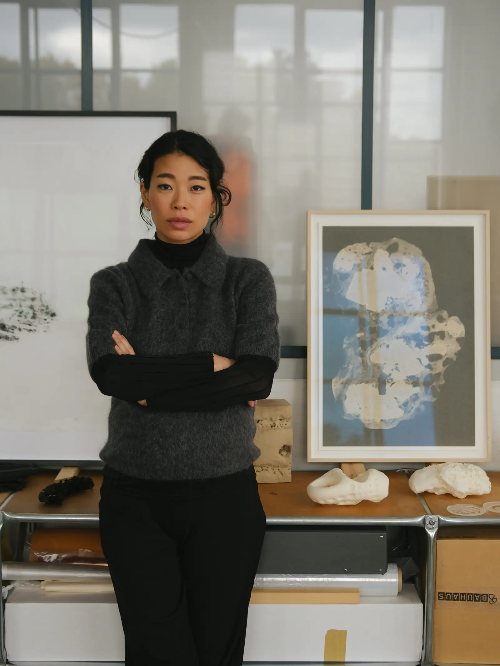 Porträtfoto der deutsch-koreanischen Künsterlin Jeewi Lee, die mit verschränkten Armen vor eigenen Werken steht.