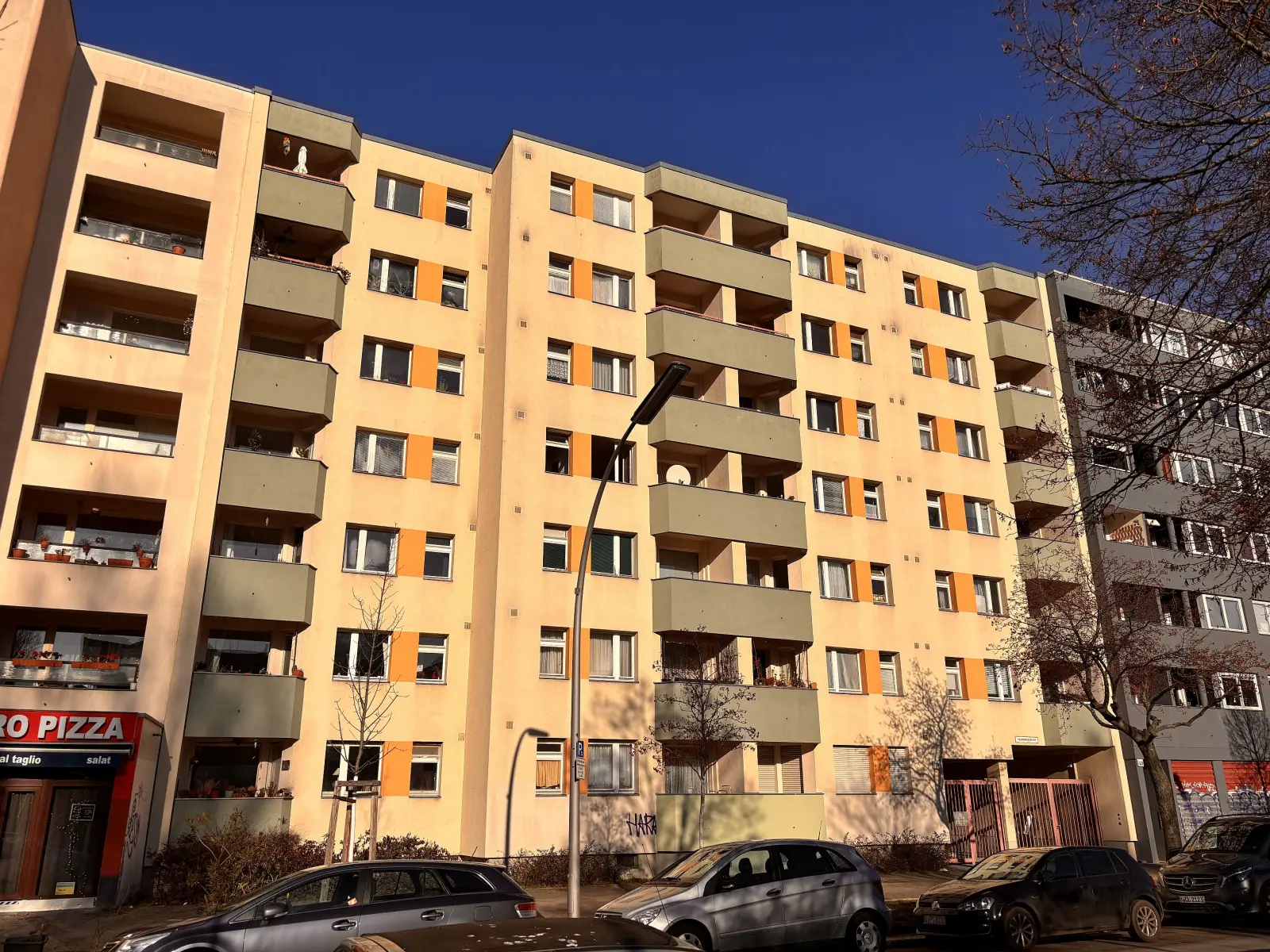Mehrgeschossiges Wohnhaus in der Berliner Winterfeldstraße mit Balkonen, parkenden Autos und Straßenraum im Sonnenlicht.