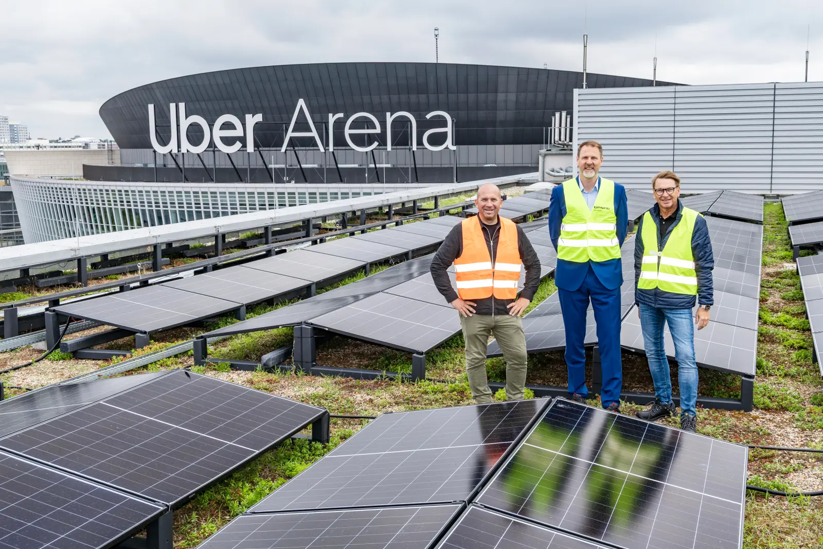 Christian Schnürle, Ole Hertel und Matthias Trunk stehen zwischen Reihen von Solarmodulen auf dem Dach der Uber Eats Music Hall in Berlin. Bild mit KI erstellt.