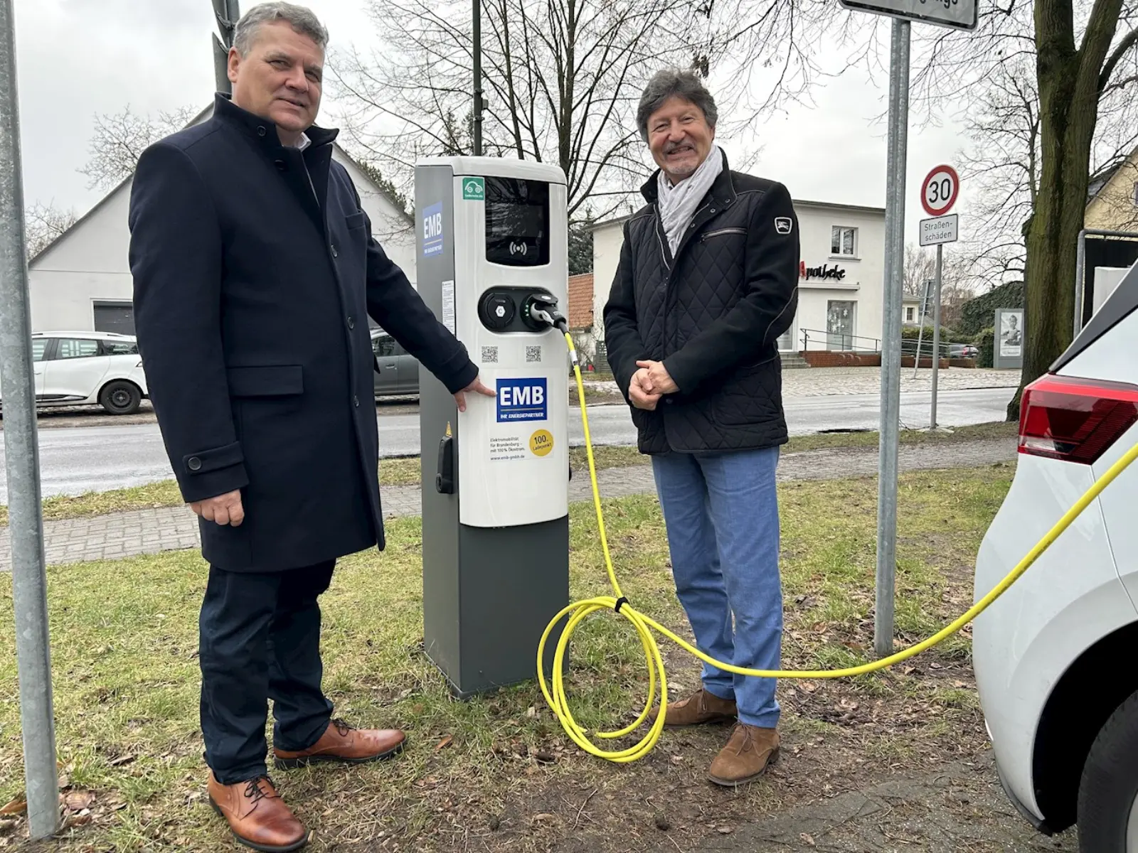 Zwei Männer mit schwarzen Jacken stehen neben einer E-Auto-Ladesäule, während ein Stromkabel dort eingesteckt ist.