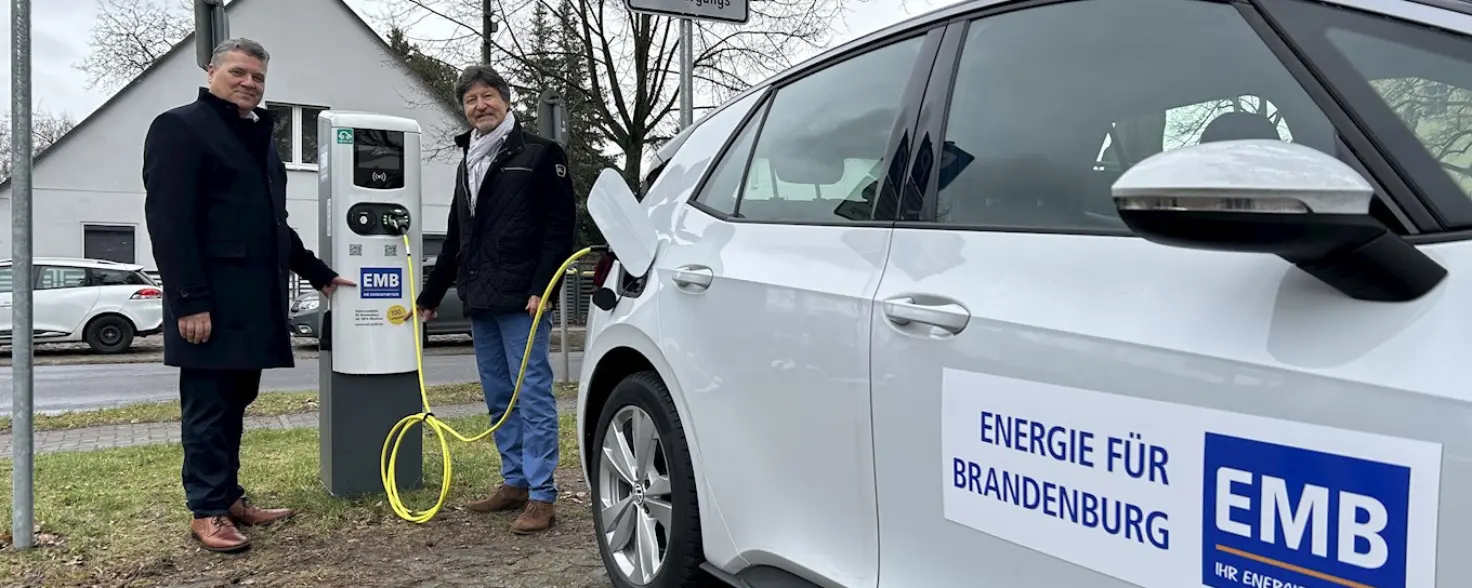 Im Vordergrund ist ein E-Auto, das an einer E-Auto-Ladesäule eingesteckt ist. Zwei Männer stehen neben der Ladesäule.