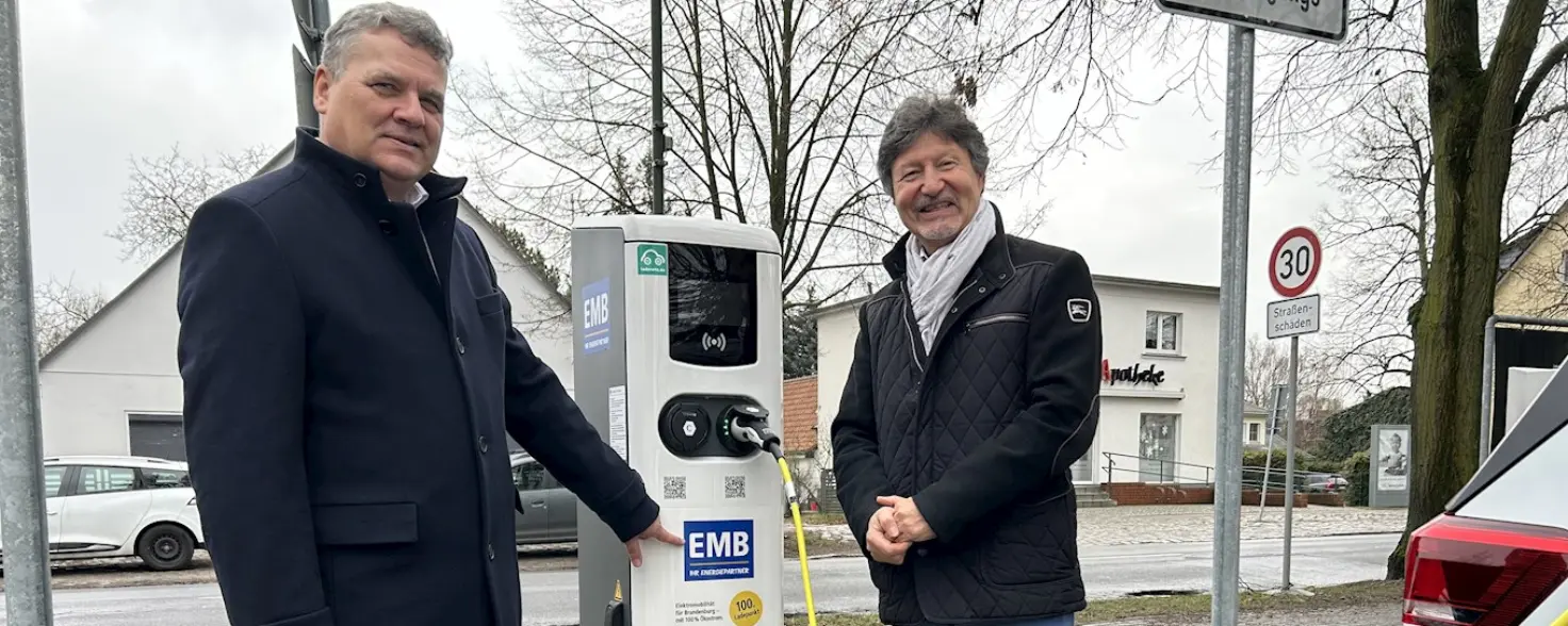Zwei Männer mit schwarzen Jacken stehen neben einer E-Auto-Ladesäule, während ein Stromkabel dort eingesteckt ist.