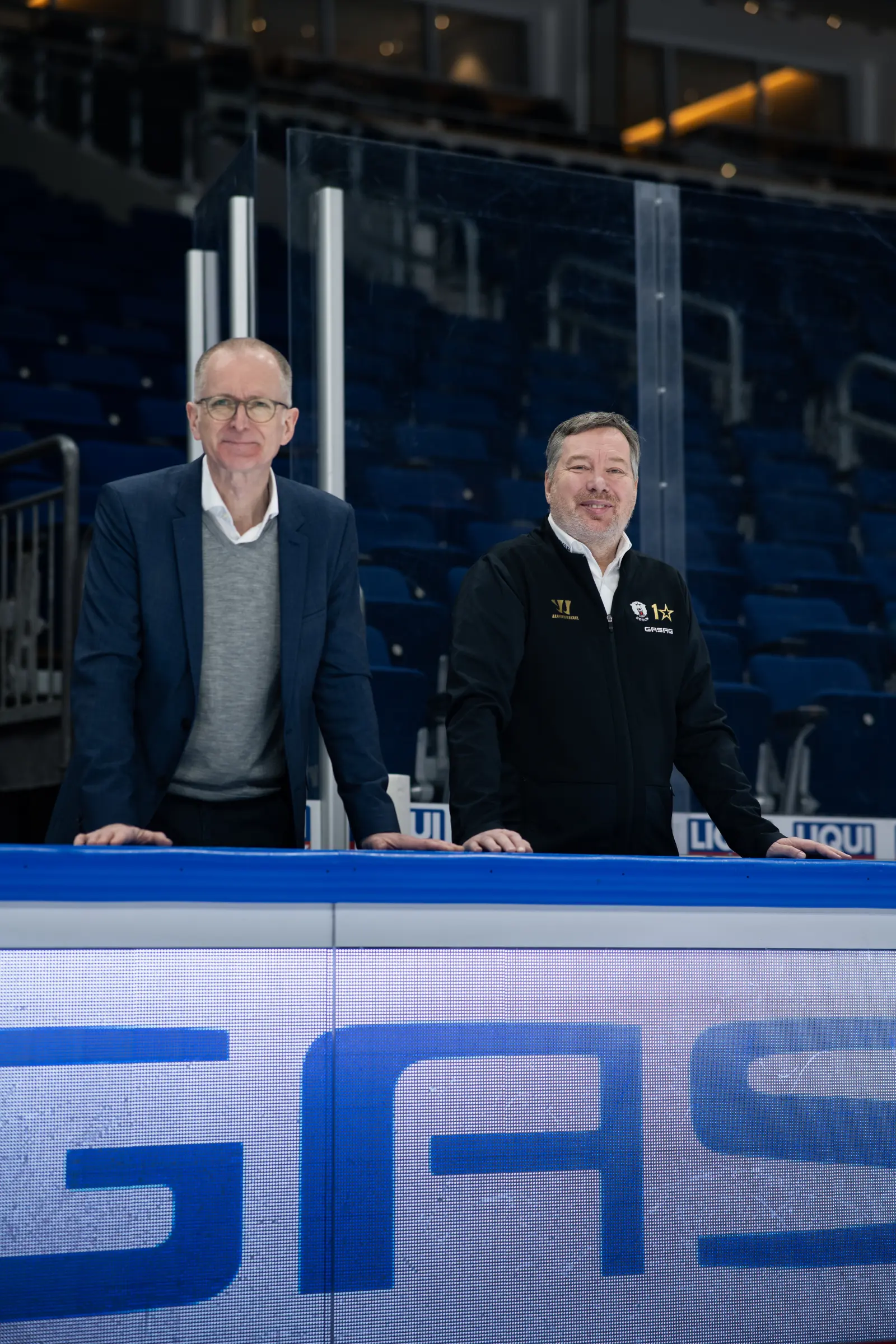 Georg Friedrichs, GASAG-Vorstandsvorsitzender, und Thomas Bothstede, Geschäftsführer der EHC Eisbären Berlin, stützen sich auf die Bande im Stadion.