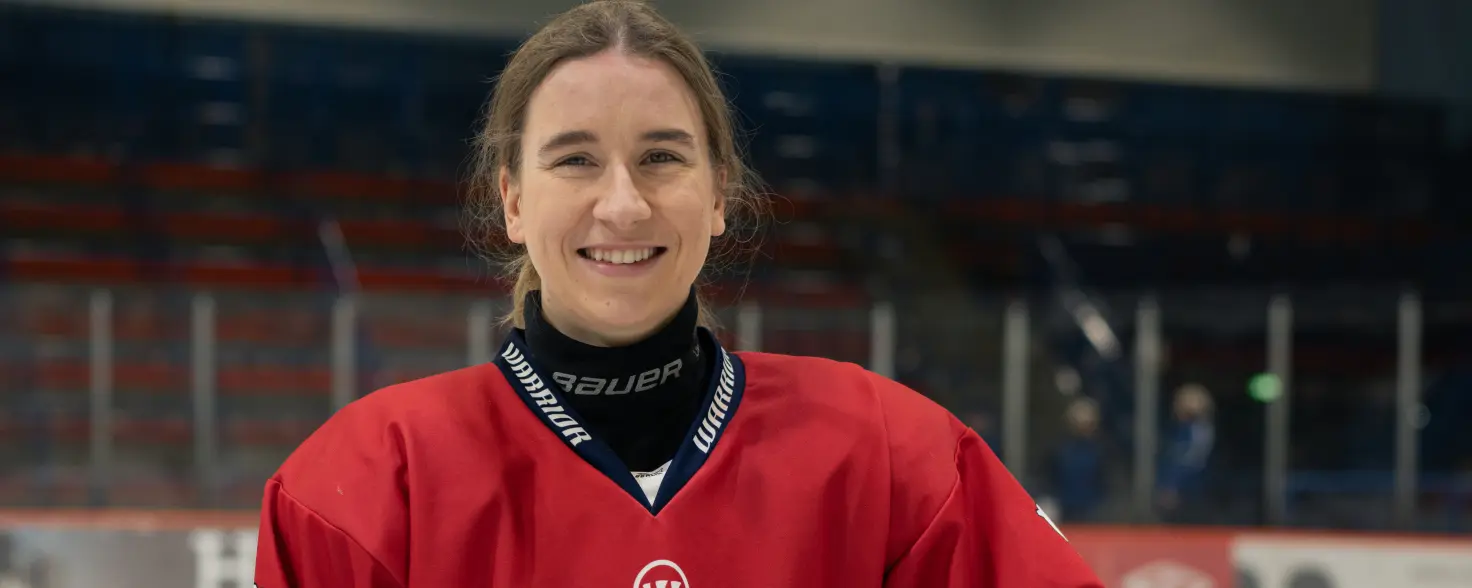 Laura Kluge von den Eisbären Juniors hat einen weißen Helm unter dem Arm und steht im roten Eishockey-Trikot vor dem Tor im Hintergrund.