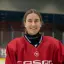 Laura Kluge von den Eisbären Juniors hat einen weißen Helm unter dem Arm und steht im roten Eishockey-Trikot vor dem Tor im Hintergrund.