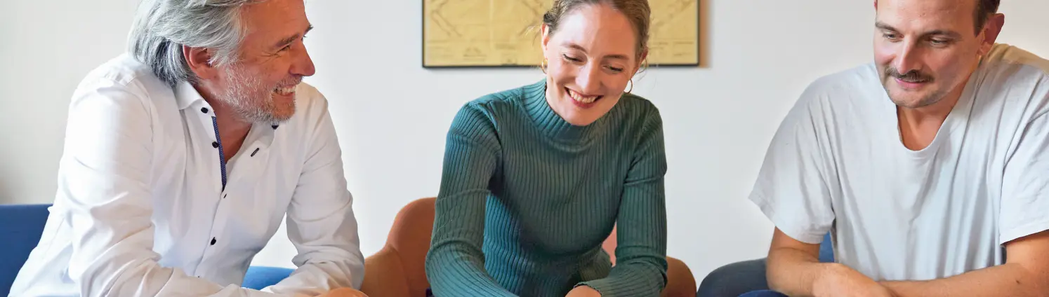 Ein Team aus einer Frau und zwei Männern am Tisch und diskutiert. Die Frau im Rollkragenpulli deutet auf einen großen Plan auf der Tischplatte.