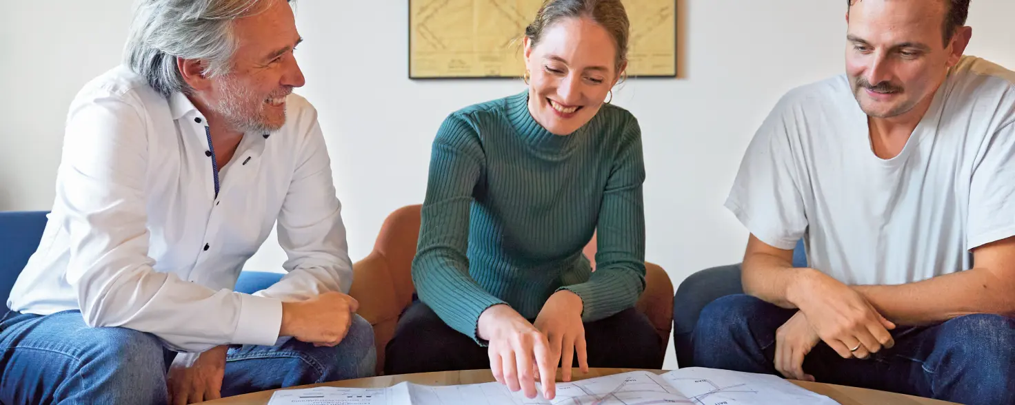 Ein Team aus einer Frau und zwei Männern am Tisch und diskutiert. Die Frau im Rollkragenpulli deutet auf einen großen Plan auf der Tischplatte.
