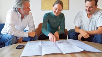Ein Team aus einer Frau und zwei Männern am Tisch und diskutiert. Die Frau im Rollkragenpulli deutet auf einen großen Plan auf der Tischplatte.